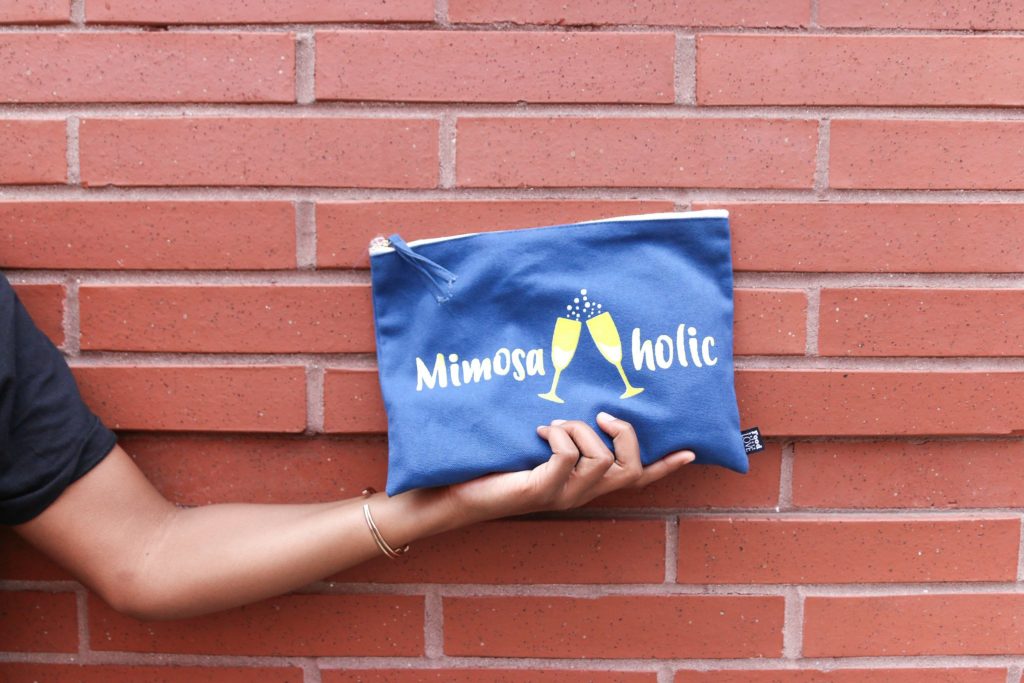 A clutch with the words "mimisaholic" on it and two clinking champagne glasses.